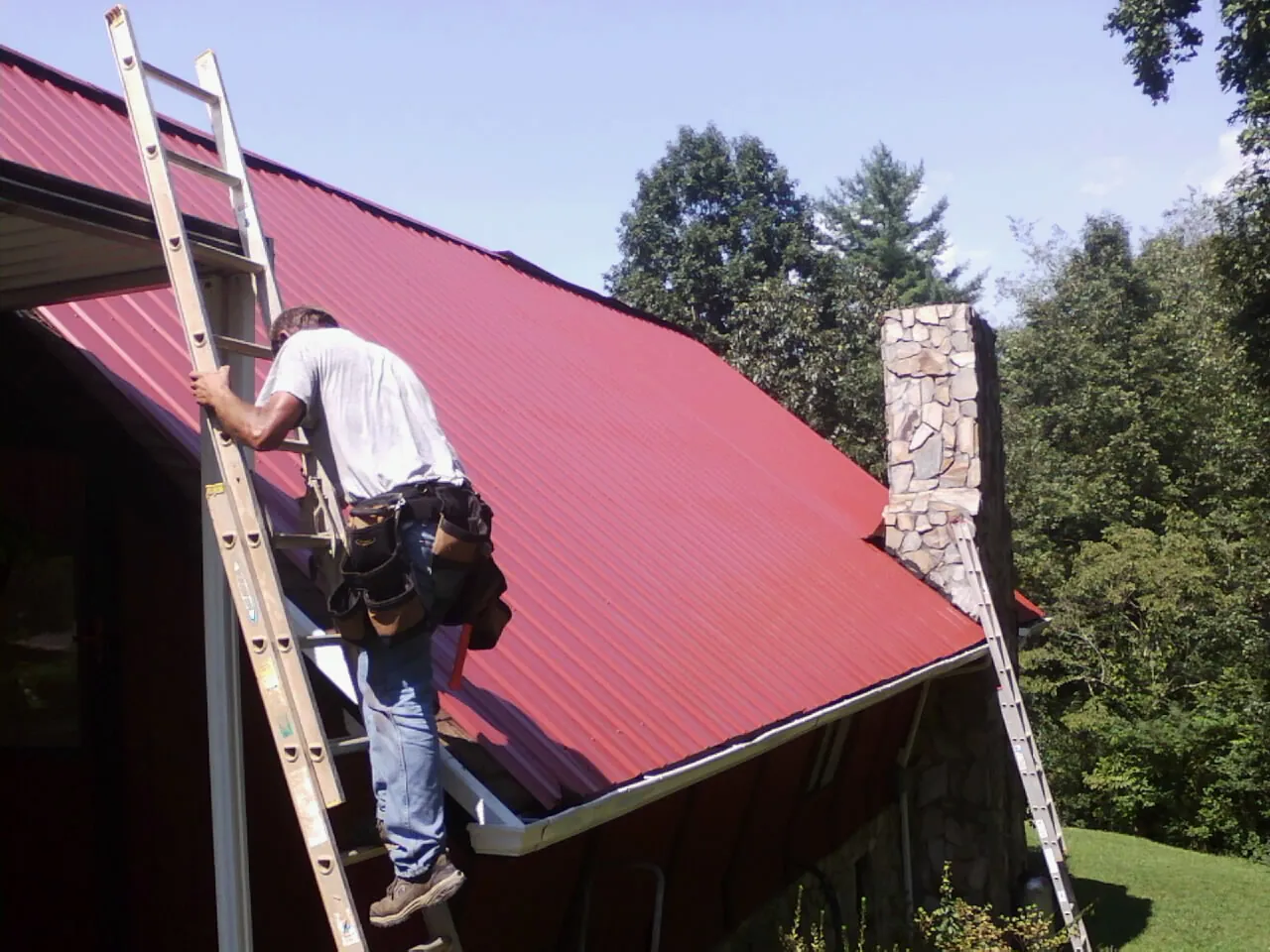 Roofer on a ladder beside a red metal roof and stone chimney — Soffit & Fascia Repair by Roofing Brothers in Leeton